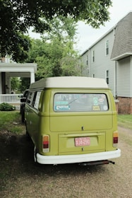 A vintage green van is parked on a grassy area beside a residential building. The vehicle features various stickers on its rear window, including messages like 'Vote Green' and 'Hippie Power'. The scene is shaded by overhanging leafy branches, and adjacent to the van is another classic vehicle.
