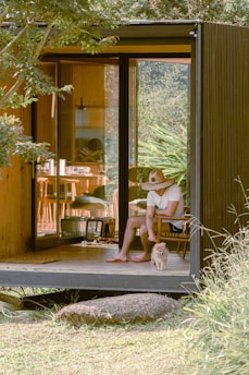 A person wearing a large straw hat is seated on a wooden chair inside a modern, cozy cabin with sliding glass doors. They are reaching down to pet a small fluffy dog standing at their feet. The interior of the cabin features wooden accents and minimalistic furniture, with greenery visible outside through the open doors. Sunlight filters through the trees, casting dappled shadows across the scene.