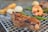 A grilled piece of meat with a slightly charred surface is placed on a metal grate, alongside roasted shrimp and green vegetables. Blurred in the background, there are various grilled items including what appears to be bread and onions.