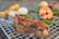 A grilled piece of meat with a slightly charred surface is placed on a metal grate, alongside roasted shrimp and green vegetables. Blurred in the background, there are various grilled items including what appears to be bread and onions.