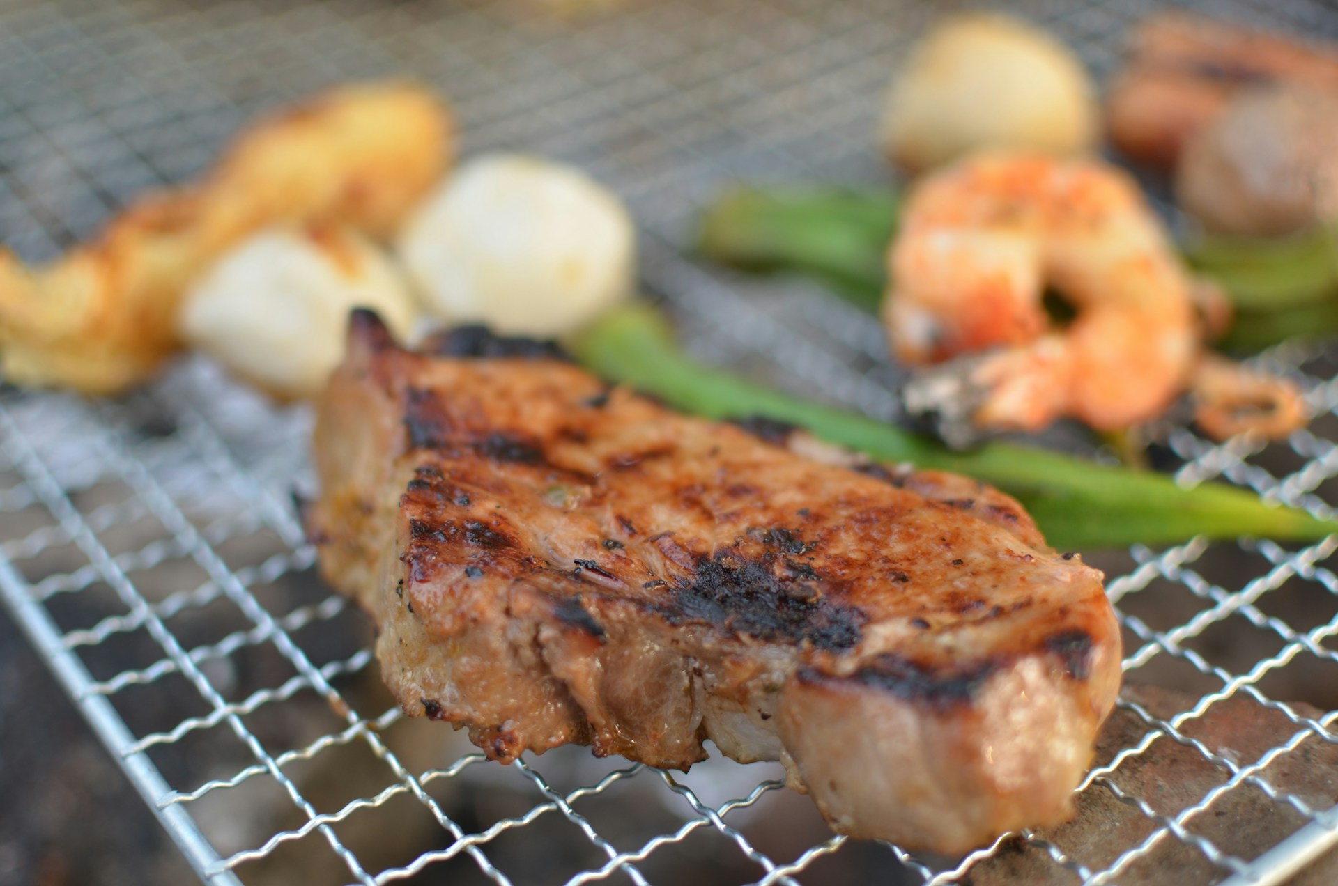 A grilled piece of meat with a slightly charred surface is placed on a metal grate, alongside roasted shrimp and green vegetables. Blurred in the background, there are various grilled items including what appears to be bread and onions.