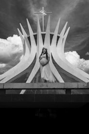 A pregnant woman in a long, flowing dress with a floral headband stands in front of a modern, abstract architectural structure with a large cross on top. The sky is partly cloudy, with dramatic cloud formations.