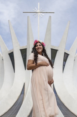 A pregnant woman wearing a flowing pink dress stands in front of a modern architectural structure with tall, white, pointed elements reaching towards the sky. She has a flower crown on her head and is smiling gently.
