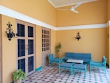 A cozy patio area with tiled flooring, consisting of wooden doors and windows. Against the light-orange walls, there is a seating arrangement of turquoise metal-framed furniture including a sofa, chairs, and a coffee table. Black wall-mounted lanterns are attached on either side of the doors, and potted plants add a touch of greenery.