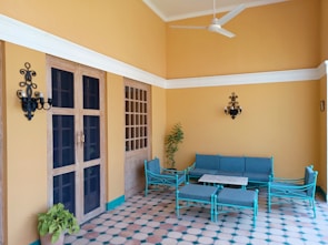 A cozy patio area with tiled flooring, consisting of wooden doors and windows. Against the light-orange walls, there is a seating arrangement of turquoise metal-framed furniture including a sofa, chairs, and a coffee table. Black wall-mounted lanterns are attached on either side of the doors, and potted plants add a touch of greenery.