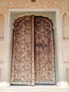 An intricately carved wooden door set within an ornately decorated stone frame. The door features geometric and floral patterns, with metal studs and handles that enhance its antique appearance. Surrounding the door are wall panels with detailed floral motifs painted in a pastel shade.