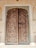 An intricately carved wooden door set within an ornately decorated stone frame. The door features geometric and floral patterns, with metal studs and handles that enhance its antique appearance. Surrounding the door are wall panels with detailed floral motifs painted in a pastel shade.