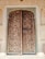 An intricately carved wooden door set within an ornately decorated stone frame. The door features geometric and floral patterns, with metal studs and handles that enhance its antique appearance. Surrounding the door are wall panels with detailed floral motifs painted in a pastel shade.