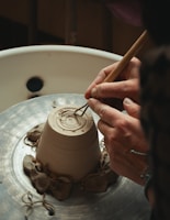 Hands are shaping a clay pot on a spinning pottery wheel, with delicate tools carving intricate patterns into the base. The setting is rustic and focused on a detailed view of the craftsmanship involved in pottery making.