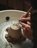 Hands are shaping a clay pot on a spinning pottery wheel, with delicate tools carving intricate patterns into the base. The setting is rustic and focused on a detailed view of the craftsmanship involved in pottery making.
