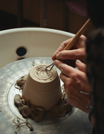 Hands are shaping a clay pot on a spinning pottery wheel, with delicate tools carving intricate patterns into the base. The setting is rustic and focused on a detailed view of the craftsmanship involved in pottery making.