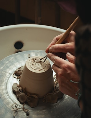 Hands are shaping a clay pot on a spinning pottery wheel, with delicate tools carving intricate patterns into the base. The setting is rustic and focused on a detailed view of the craftsmanship involved in pottery making.