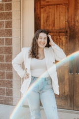 A woman with long, curly hair is smiling and posing in front of a wooden door. She is wearing a white blazer, a light colored top, and jeans. A rainbow light effect crosses diagonally in front of her.