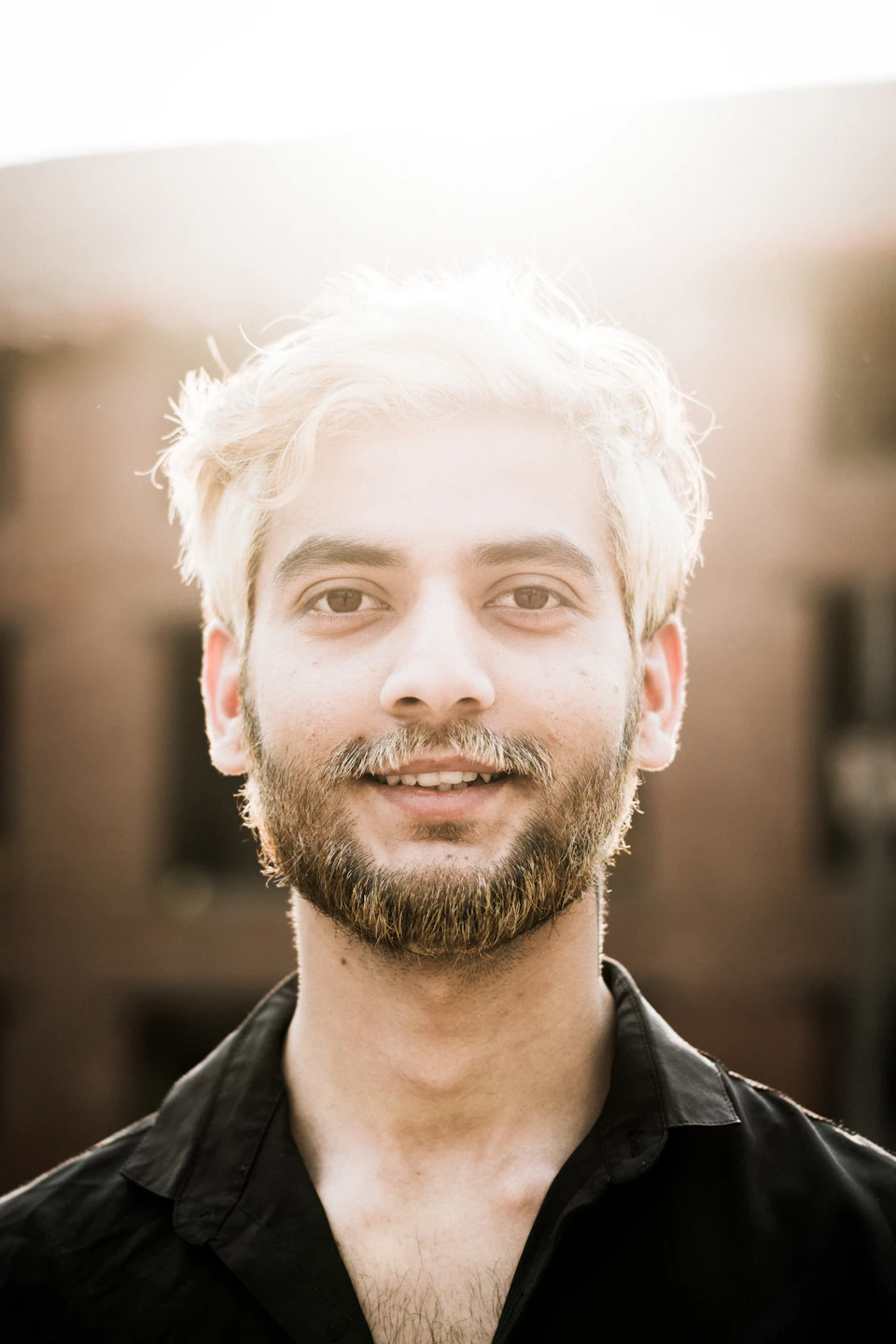 Young man with blonde hair and beard stands against a brick wall, smiling directly