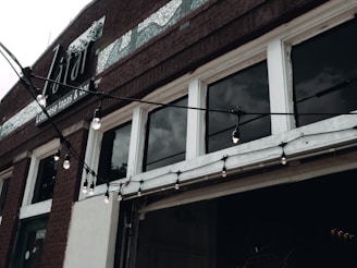The facade of a building is shown with large windows and a brick exterior. The windows are separated by white frames and are slightly reflective, showing a cloudy sky. Hanging string lights with exposed bulbs are draped across the building just above the entrance. Above the windows, there is signage indicating it is a Lebanese tapas and bar.