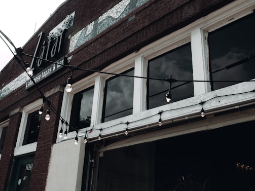 The facade of a building is shown with large windows and a brick exterior. The windows are separated by white frames and are slightly reflective, showing a cloudy sky. Hanging string lights with exposed bulbs are draped across the building just above the entrance. Above the windows, there is signage indicating it is a Lebanese tapas and bar.