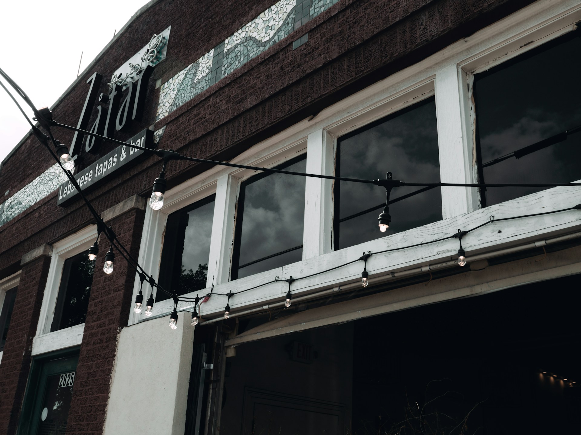 The facade of a building is shown with large windows and a brick exterior. The windows are separated by white frames and are slightly reflective, showing a cloudy sky. Hanging string lights with exposed bulbs are draped across the building just above the entrance. Above the windows, there is signage indicating it is a Lebanese tapas and bar.