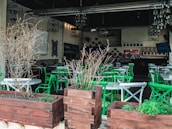 An empty restaurant patio featuring green and white chairs arranged around circular tables. Wooden planters with dry branches are placed in the foreground, and chandeliers hang from the ceiling. The interior is dimly lit, showcasing decorative elements on the walls and shelves with bottles in the background.