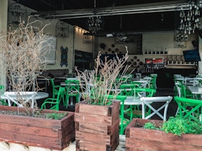 An empty restaurant patio featuring green and white chairs arranged around circular tables. Wooden planters with dry branches are placed in the foreground, and chandeliers hang from the ceiling. The interior is dimly lit, showcasing decorative elements on the walls and shelves with bottles in the background.