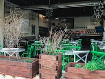 An empty restaurant patio featuring green and white chairs arranged around circular tables. Wooden planters with dry branches are placed in the foreground, and chandeliers hang from the ceiling. The interior is dimly lit, showcasing decorative elements on the walls and shelves with bottles in the background.