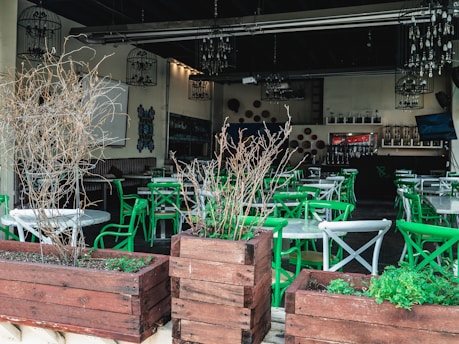 An empty restaurant patio featuring green and white chairs arranged around circular tables. Wooden planters with dry branches are placed in the foreground, and chandeliers hang from the ceiling. The interior is dimly lit, showcasing decorative elements on the walls and shelves with bottles in the background.