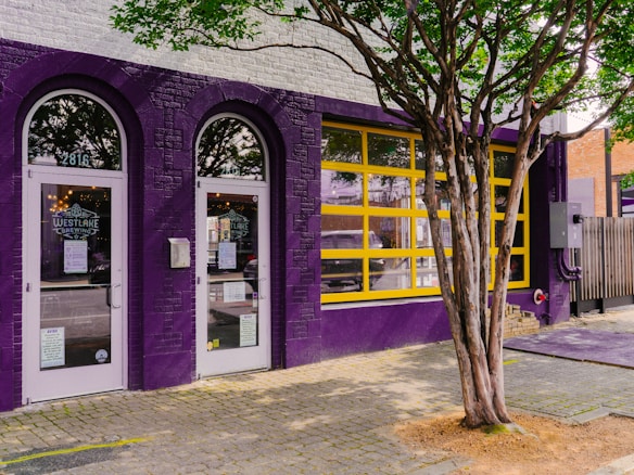 A storefront with vibrant purple brick walls and yellow-framed windows. The store has two glass doors with the logo and name 'Westlake Brewing' displayed. A large tree with a twisted trunk is in the foreground on a paved sidewalk. Inside, some lights are visible through the glass.
