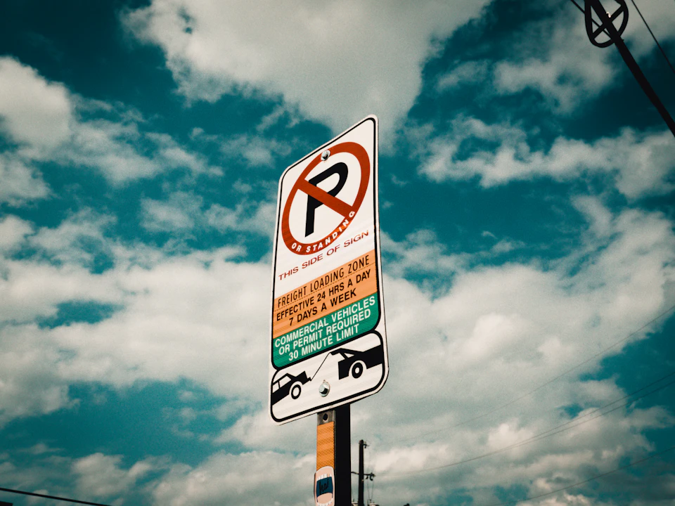 The image shows a street sign mounted on a pole, displaying a "No Parking" restriction. The sign is white with black text and features an orange circle with a red border around it, indicating that parking is not allowed in the designated zone. Below the main sign, there are two smaller signs providing additional information about the parking restrictions: one stating "No Parking Zone", and another stating "Fishing Zone".