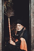 An elderly man is seated in a dimly lit doorway, holding a decorated staff. He wears glasses, a turban, and traditional attire featuring an orange scarf. The setting suggests a rustic or spiritual atmosphere.