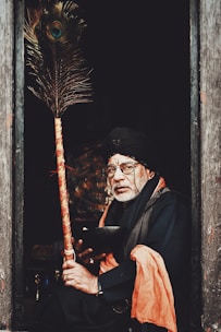An elderly man is seated in a dimly lit doorway, holding a decorated staff. He wears glasses, a turban, and traditional attire featuring an orange scarf. The setting suggests a rustic or spiritual atmosphere.