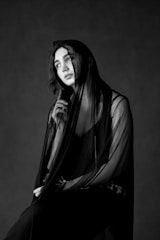 A person is seated in a thoughtful pose, draped in a sheer, dark fabric. The monochrome setting accentuates the soft lighting and contrasts, highlighting the contemplative expression.