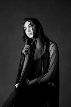 A person is seated in a thoughtful pose, draped in a sheer, dark fabric. The monochrome setting accentuates the soft lighting and contrasts, highlighting the contemplative expression.