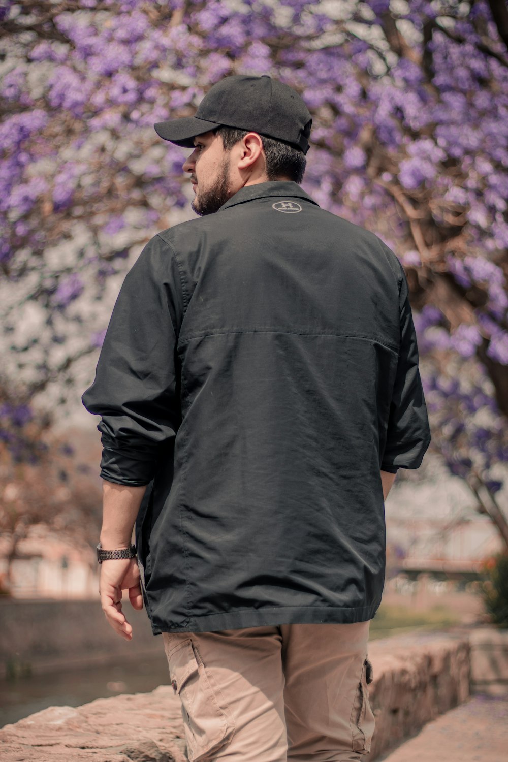 man in black polo shirt and black hat standing near purple flowers during daytime