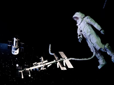 An astronaut in a space suit is floating in the dark environment of outer space, tethered to a spacecraft. A satellite is visible to the left, while the International Space Station can be seen in the background, illuminated against the blackness.