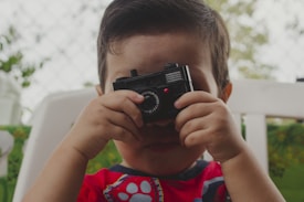 A child is holding a small black camera to their face as if taking a picture. The child is seated outdoors on a white chair, wearing a red shirt with a colorful design on the front. The background features blurred greenery and what appears to be a fence.