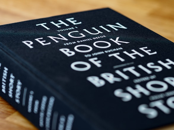 A close-up view of a hardcover book titled 'The Penguin Book of the British Short Story'. The book cover is black with white and teal text. It is placed on a wooden surface, slightly tilted to the right.