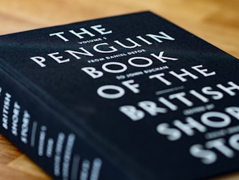 A close-up view of a hardcover book titled 'The Penguin Book of the British Short Story'. The book cover is black with white and teal text. It is placed on a wooden surface, slightly tilted to the right.