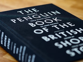 A close-up view of a hardcover book titled 'The Penguin Book of the British Short Story'. The book cover is black with white and teal text. It is placed on a wooden surface, slightly tilted to the right.