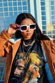 A woman wearing oversized white sunglasses and a brown jacket stands confidently. Her T-shirt features a vivid image of a man with dreadlocks blending into a lion's mane. She is accessorized with a chain necklace and has her hand resting on her sunglasses.