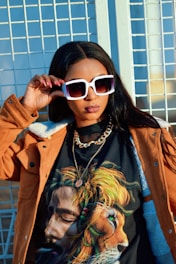 A woman wearing oversized white sunglasses and a brown jacket stands confidently. Her T-shirt features a vivid image of a man with dreadlocks blending into a lion's mane. She is accessorized with a chain necklace and has her hand resting on her sunglasses.