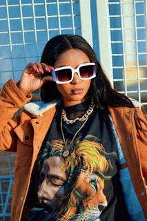 A woman wearing oversized white sunglasses and a brown jacket stands confidently. Her T-shirt features a vivid image of a man with dreadlocks blending into a lion's mane. She is accessorized with a chain necklace and has her hand resting on her sunglasses.