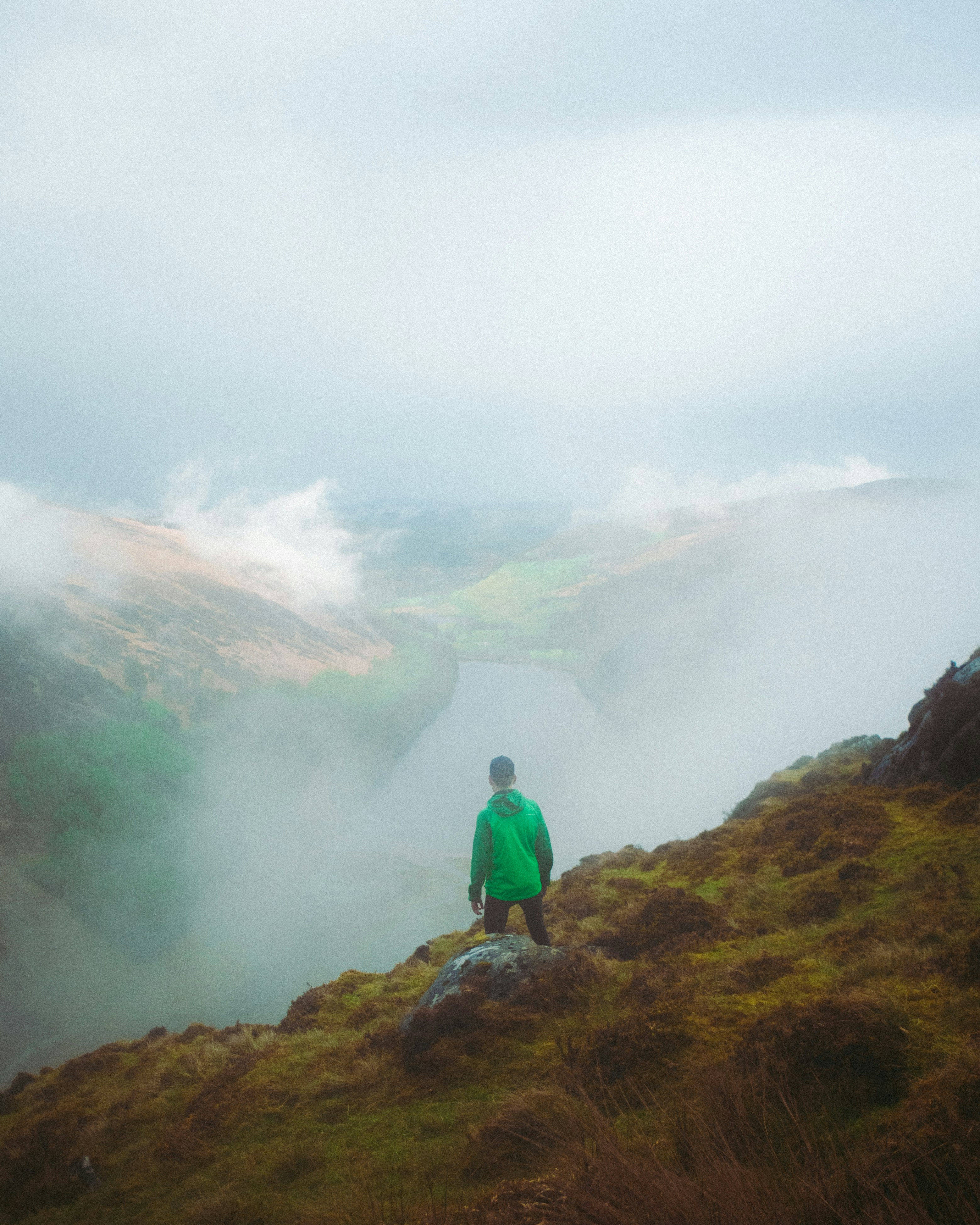 A person in a bright green jacket stands on a foggy hillside overlooking a mist-filled valley.