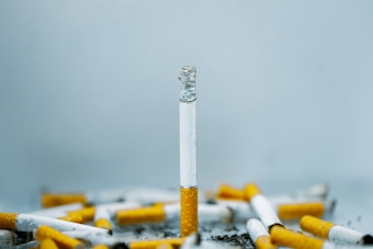 A single cigarette stands upright, surrounded by numerous cigarette butts lying scattered on a surface, creating a sense of clutter and disorder. The background is a soft, muted blue-gray, highlighting the cigarettes and their burnt tips.