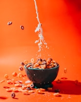 A stream of milk is being poured into a bowl filled with colorful cereal pieces, some of which are scattering around the bowl. The vibrant orange background creates a striking contrast with the white milk and the multicolored cereal.