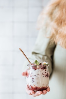 A person holds a small glass jar filled with layered yogurt, granola, and red fruit compote, topped with fresh mint leaves and small white flowers. The jar is held by a hand with manicured nails, and a gold spoon is inserted into the yogurt. The background is softly blurred, focusing attention on the jar.