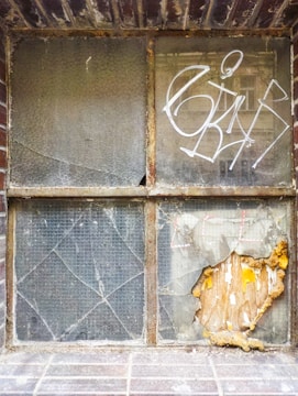 A weathered window with cracked and broken glass panes is set within a brick wall. There is graffiti in white on one of the panes and plaster with peeling paint on another, revealing orange and yellow hues underneath. The frames are rusted and there are signs of decay.