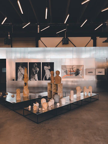 An art gallery featuring a collection of sculptures and paintings. The sculptures are displayed on black platforms and include abstract forms and figures. The wall in the background is lined with artworks, including black and white portraits and a large abstract painting with orange and grey elements. The ceiling has modern linear lights, creating a contemporary ambiance.