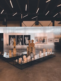 An art gallery featuring a collection of sculptures and paintings. The sculptures are displayed on black platforms and include abstract forms and figures. The wall in the background is lined with artworks, including black and white portraits and a large abstract painting with orange and grey elements. The ceiling has modern linear lights, creating a contemporary ambiance.