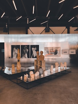 An art gallery featuring a collection of sculptures and paintings. The sculptures are displayed on black platforms and include abstract forms and figures. The wall in the background is lined with artworks, including black and white portraits and a large abstract painting with orange and grey elements. The ceiling has modern linear lights, creating a contemporary ambiance.