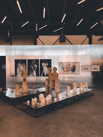 An art gallery featuring a collection of sculptures and paintings. The sculptures are displayed on black platforms and include abstract forms and figures. The wall in the background is lined with artworks, including black and white portraits and a large abstract painting with orange and grey elements. The ceiling has modern linear lights, creating a contemporary ambiance.
