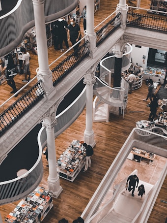 A multi-level bookstore features wooden floors, elegant white pillars, and winding staircases. People are browsing through books placed on neatly arranged tables and shelves. The architecture combines classic and modern elements, with open spaces and artistic railings.
